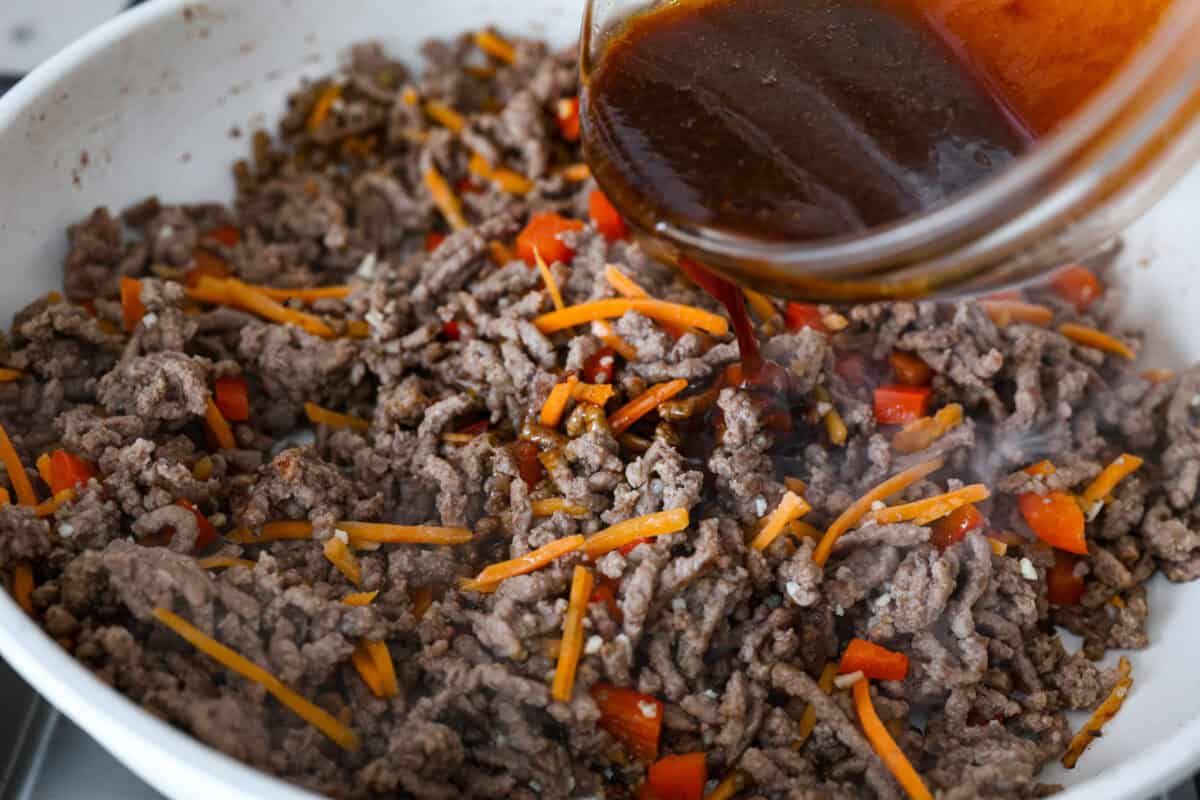 Enjoy delicious Asian flavors in these Korean Beef Lettuce Wraps! This appetizer or main is loaded with tasty lean ground beef, sautรฉed shredded carrots, red bell pepper, and garlic, all tossed in a mouthwatering sauce and spooned into lettuce leaves. Angle shot of sauce being poured over meat and vegetable mixture.
