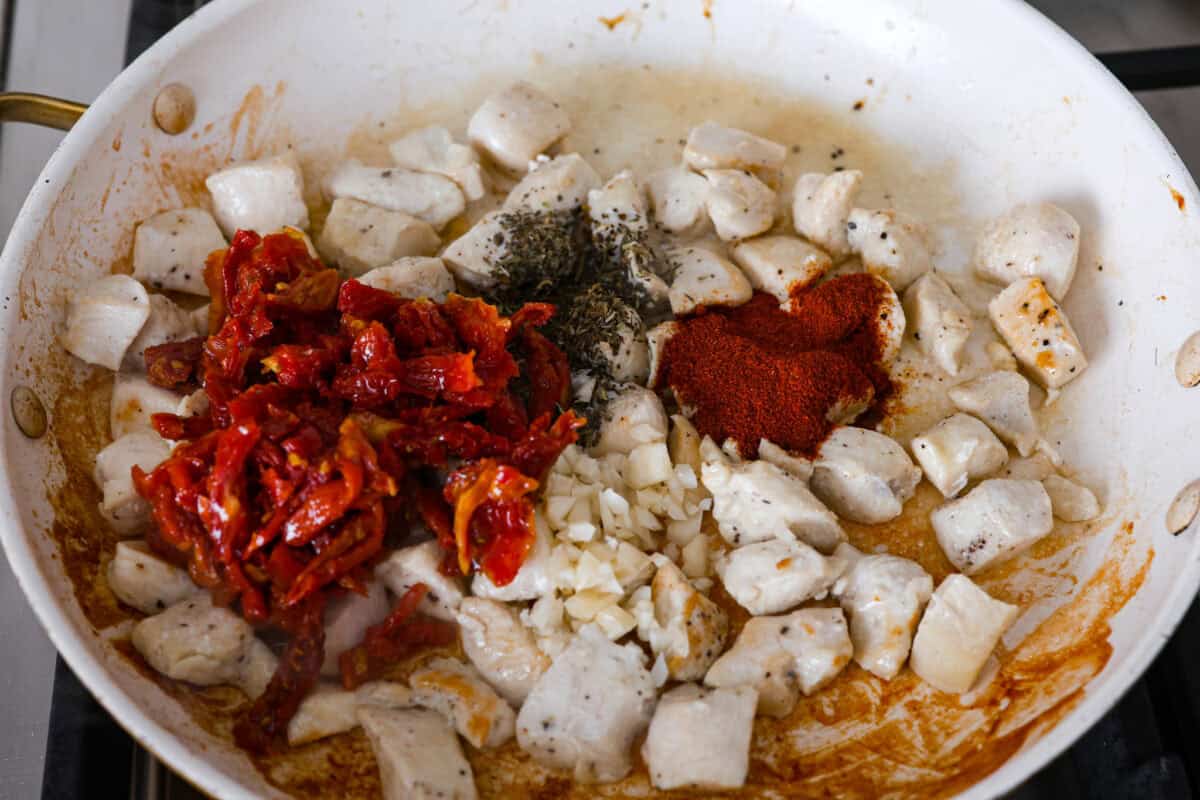 Win everyone over with One-Pan Marry Me Chicken Orzo! The recipe features tender chicken breasts cooked with orzo pasta in the most irresistible, creamy, sun-dried tomato sauce. It's quick to make and never disappoints! Overhead shot of sun-dried tomatoes, garlic and seasonings being added to the chicken cooking in a skillet.