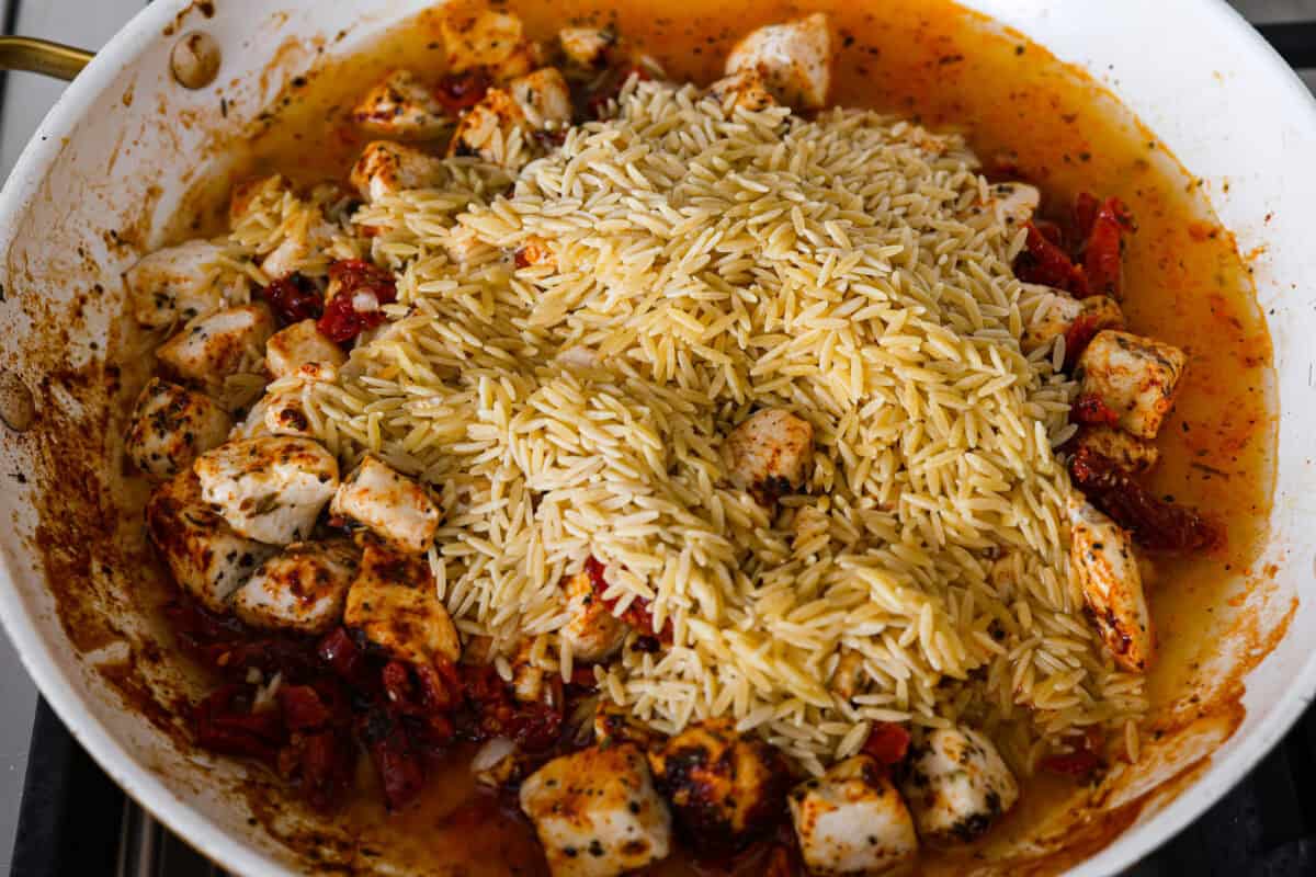 Win everyone over with One-Pan Marry Me Chicken Orzo! The recipe features tender chicken breasts cooked with orzo pasta in the most irresistible, creamy, sun-dried tomato sauce. It's quick to make and never disappoints! Overhead shot of orzo pasta being added to the chicken mixture in the skillet.