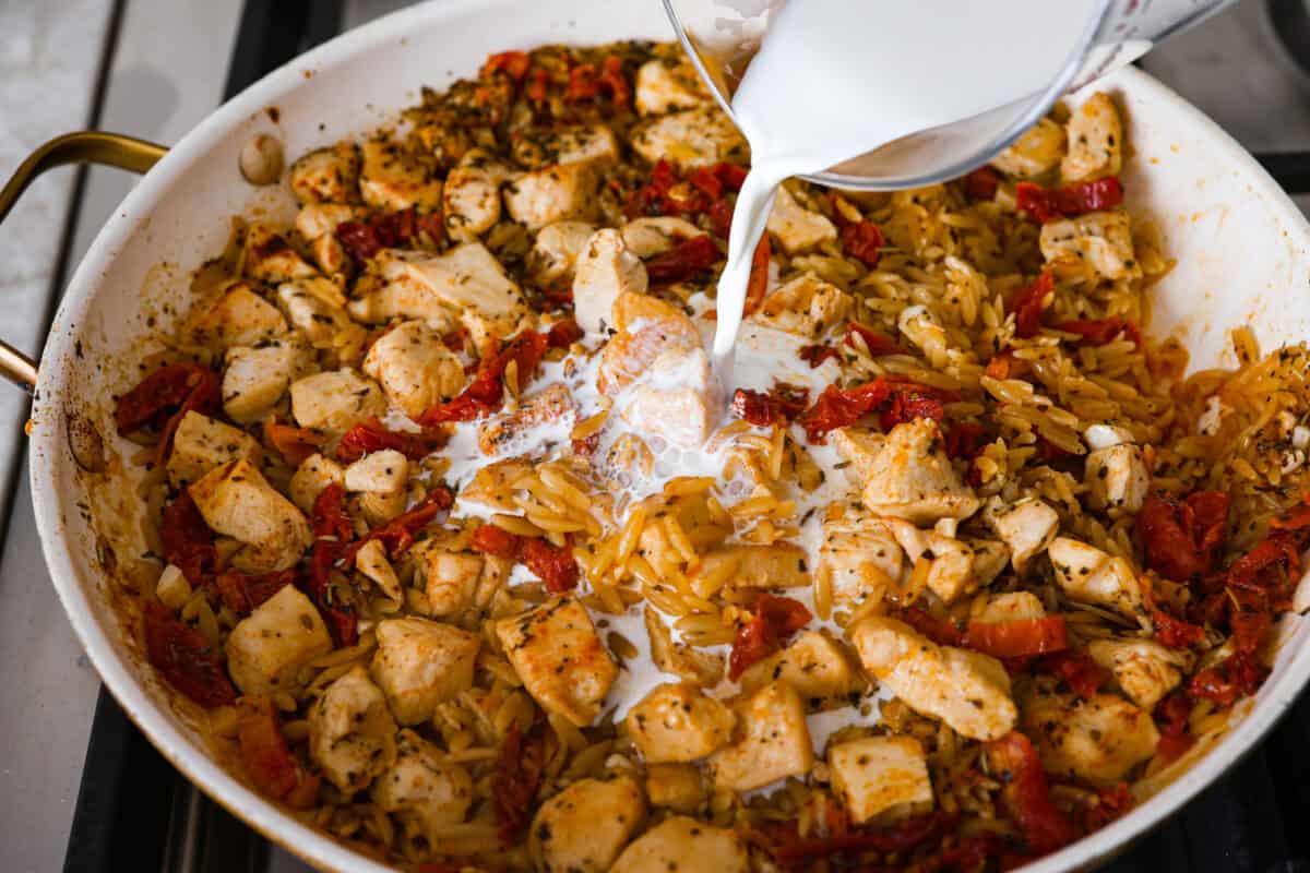 Win everyone over with One-Pan Marry Me Chicken Orzo! The recipe features tender chicken breasts cooked with orzo pasta in the most irresistible, creamy, sun-dried tomato sauce. It's quick to make and never disappoints! Overhead shot of the half-and-half being poured into the chicken mixture in the skillet.