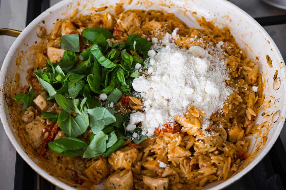 Win everyone over with One-Pan Marry Me Chicken Orzo! The recipe features tender chicken breasts cooked with orzo pasta in the most irresistible, creamy, sun-dried tomato sauce. It's quick to make and never disappoints! Overhead shot of basil and parmesan cheese being added to the skillet with chicken and orzo mixture.
