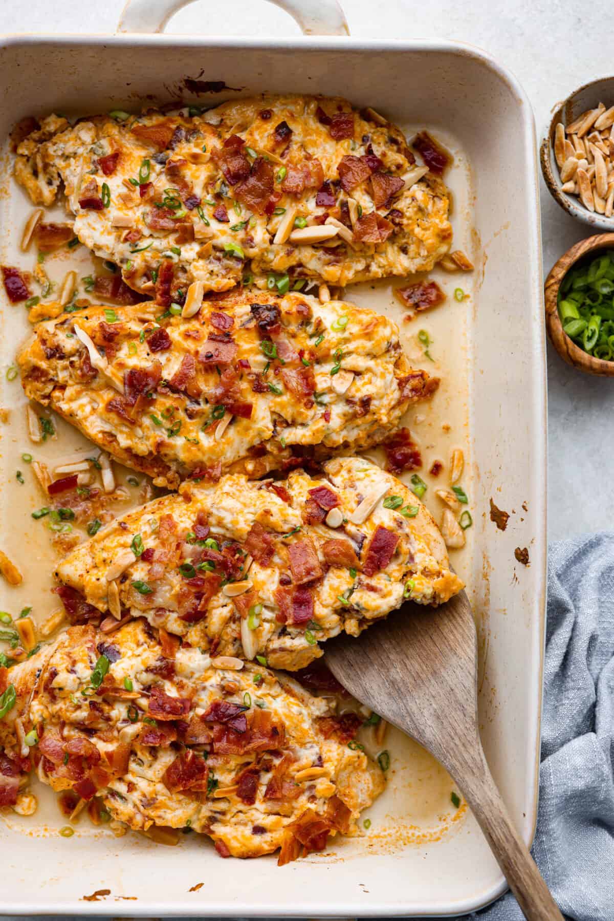 Million Dollar Chicken features juicy baked chicken topped with a creamy, cheesy layer that’s bursting with flavor. The crispy bacon and crunchy almonds really take this to the next level. Enjoy with rice, pasta, vegetables, or any of your favorite sides! Overhead shot of million dollar chicken in a baking dish with wooden serving spoon.