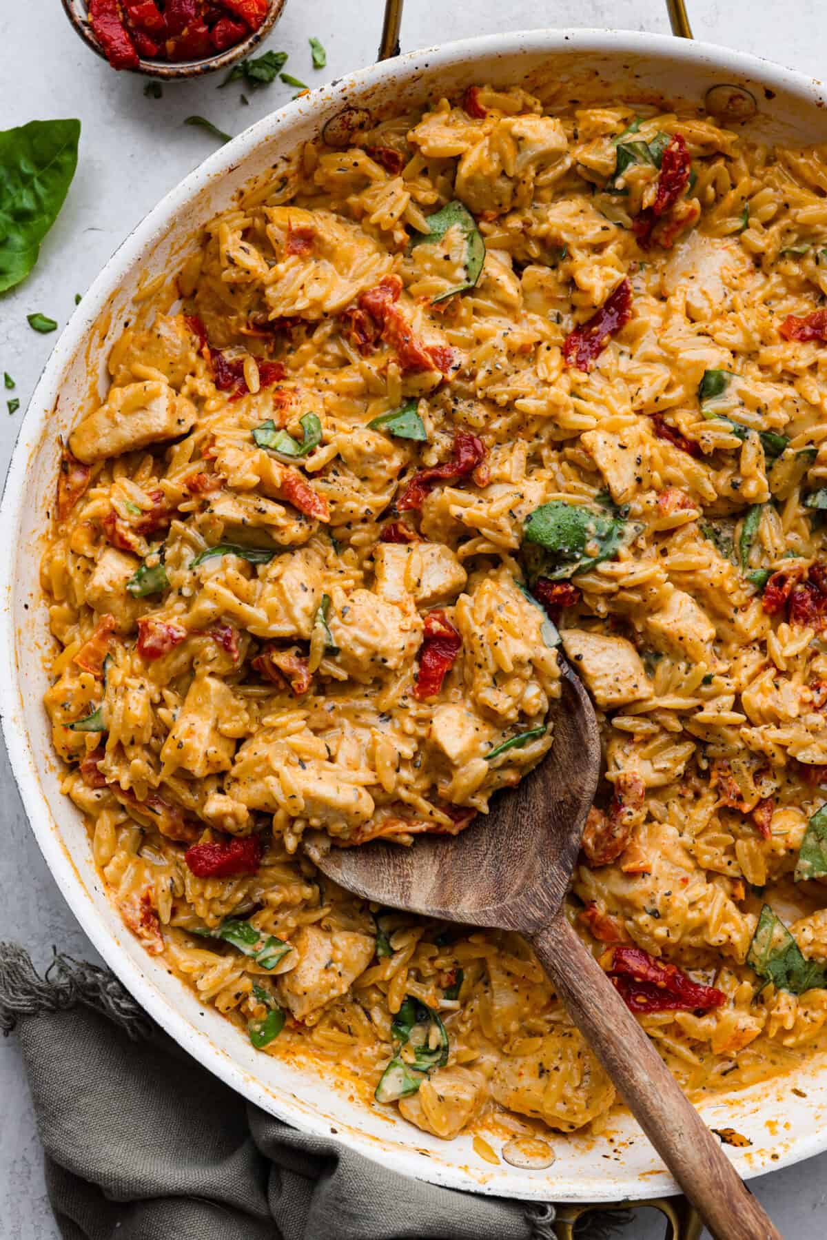 Win everyone over with One-Pan Marry Me Chicken Orzo! The recipe features tender chicken breasts cooked with orzo pasta in the most irresistible, creamy, sun-dried tomato sauce. It's quick to make and never disappoints! Overhead shot of marry me chicken orzo in a skillet with wooden serving spoon.