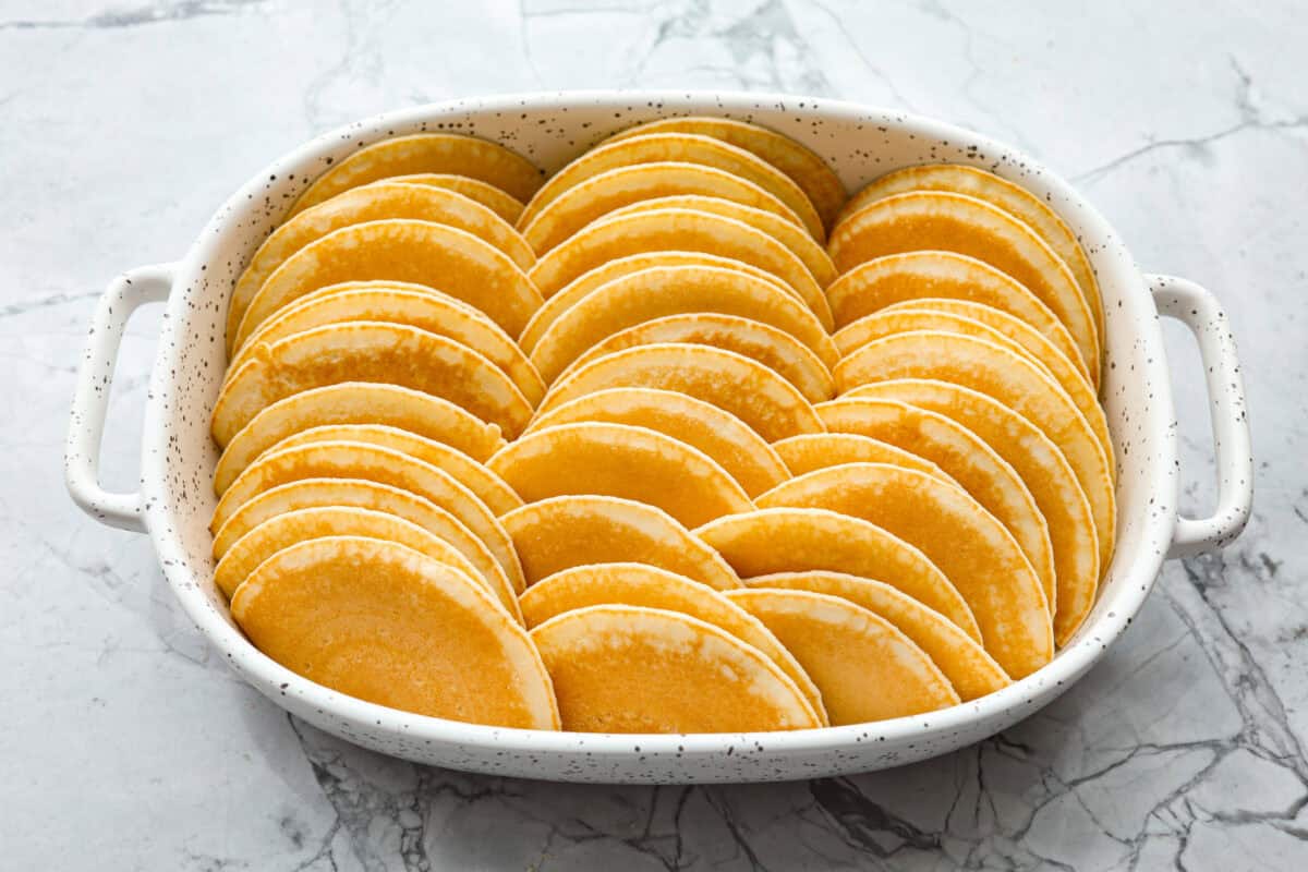 Pancake Casserole is a fun new Christmas or Easter breakfast idea. It has lots of buttery pancakes soaked overnight in a creamy custard, is baked to perfection, and served with your favorite toppings! Angle shot of pancakes stacked in baking dish.