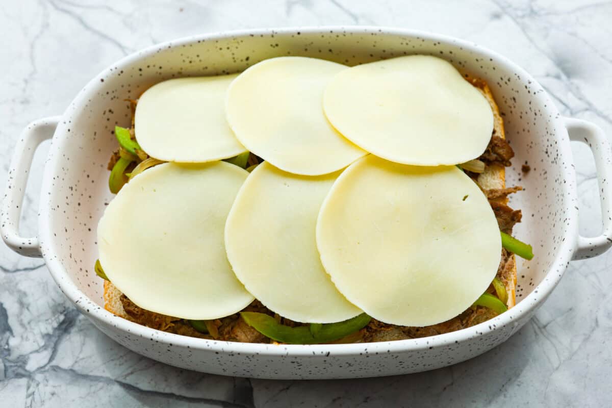 Philly cheesesteak sliders are a fun and delicious twist on a classic. These Philly cheesesteaks are filled with thinly sliced steak, caramelized onions, crisp peppers, and, of course, melty cheese. Overhead shot of slices of provolone over the veggies and beef.