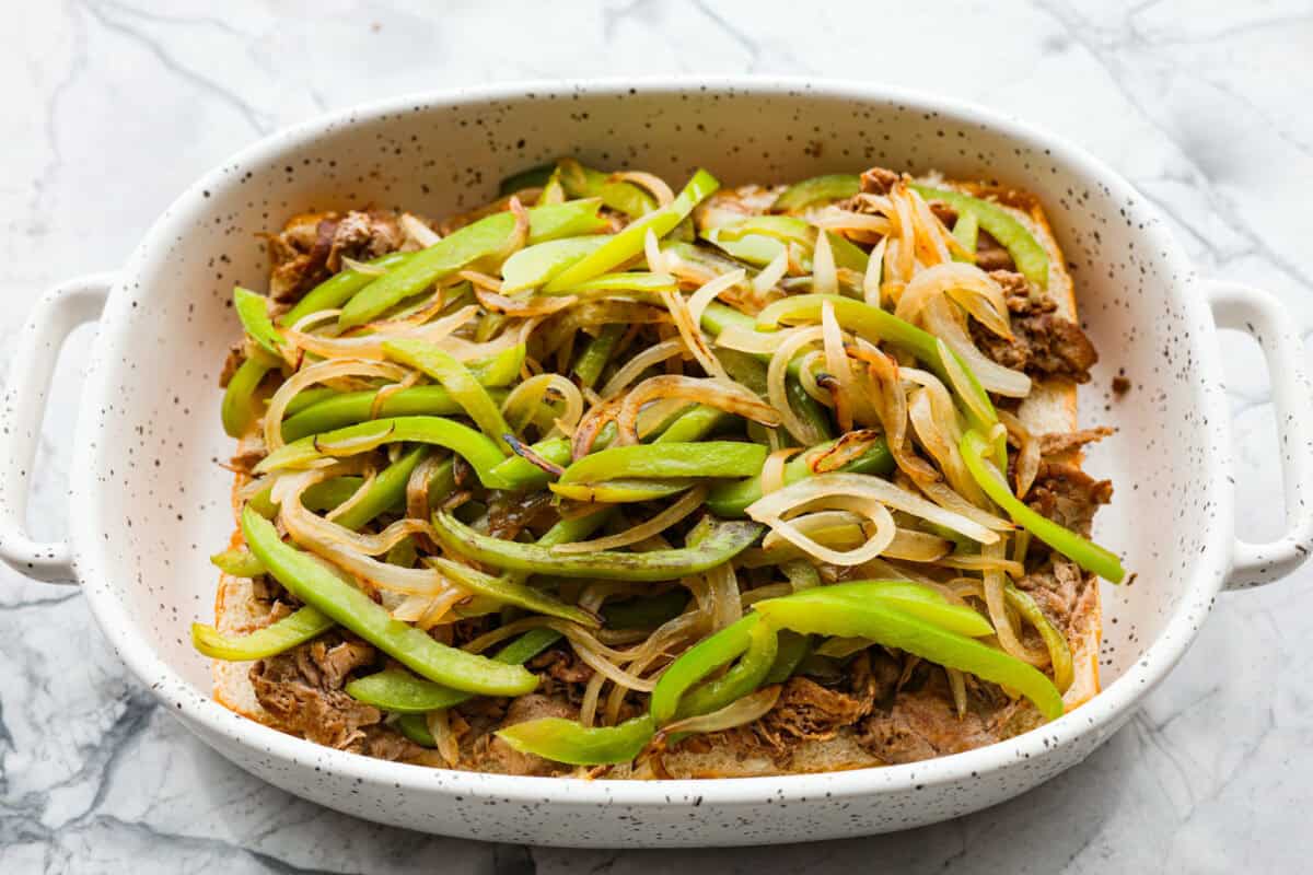 Philly cheesesteak sliders are a fun and delicious twist on a classic. These Philly cheesesteaks are filled with thinly sliced steak, caramelized onions, crisp peppers, and, of course, melty cheese. Overhead shot of pepper and onion mixture over the top of the beef in the baking dish.
