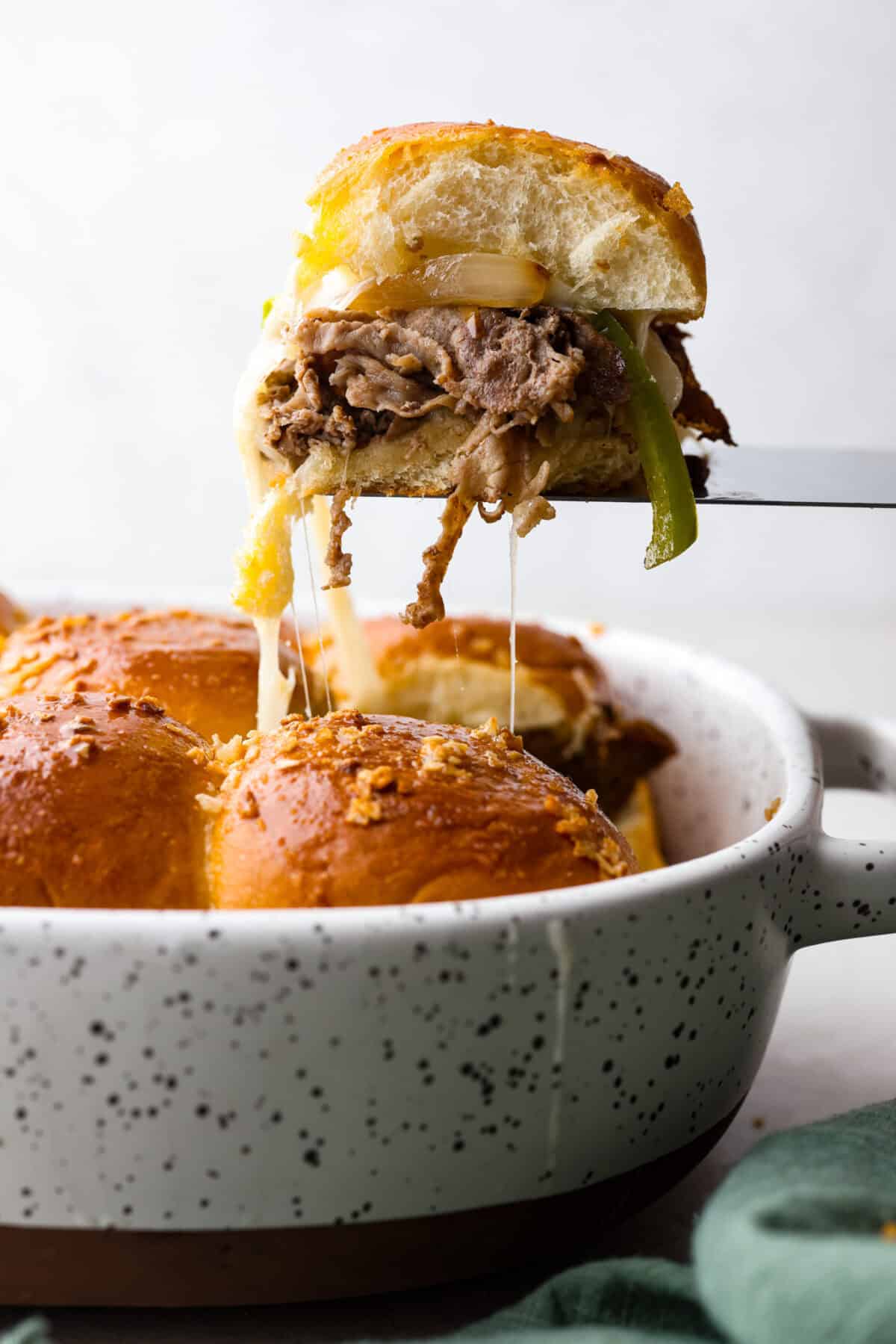 Philly cheesesteak sliders are a fun and delicious twist on a classic. These Philly cheesesteaks are filled with thinly sliced steak, caramelized onions, crisp peppers, and, of course, melty cheese. Side shot of a Philly cheesesteak slider being lifted out of the pan.