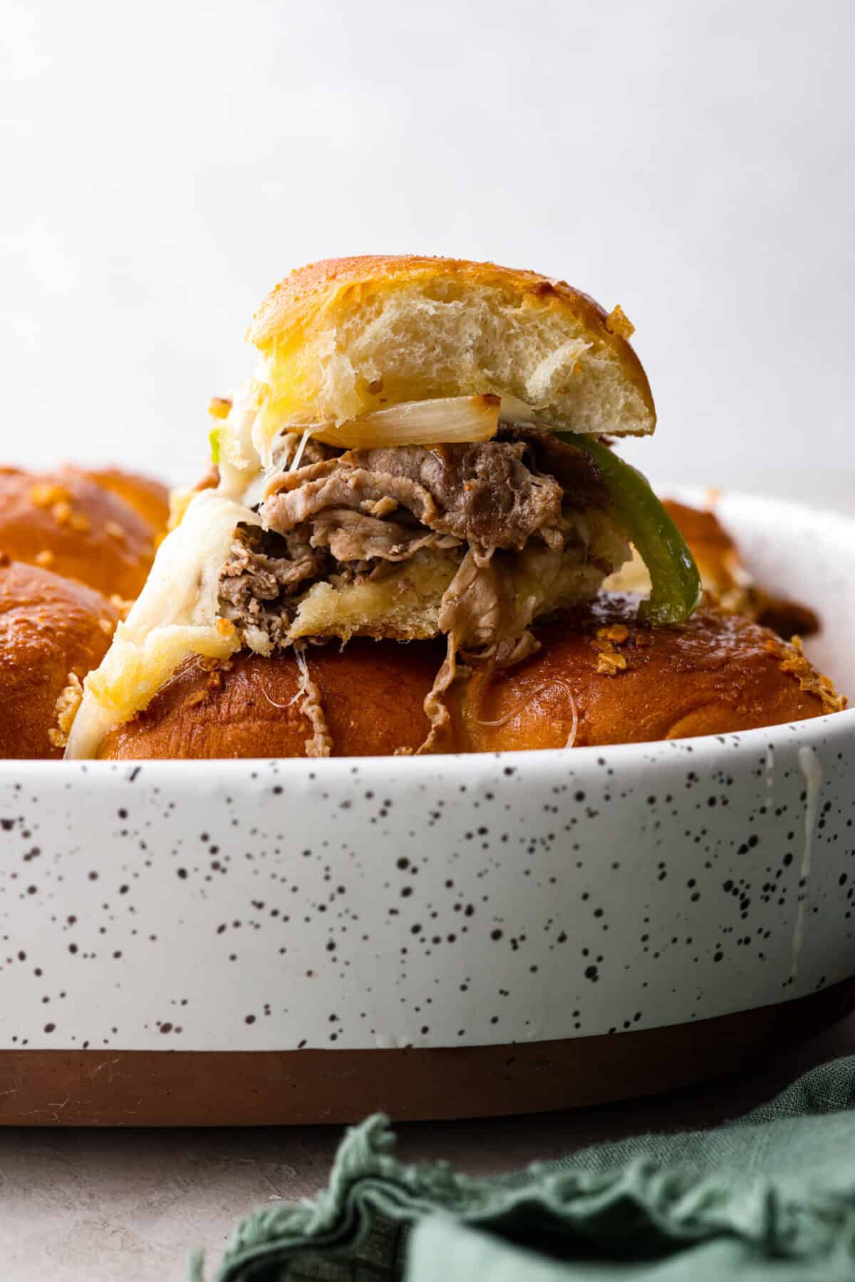 Philly cheesesteak sliders are a fun and delicious twist on a classic. These Philly cheesesteaks are filled with thinly sliced steak, caramelized onions, crisp peppers, and, of course, melty cheese. Side shot of a Philly cheesesteak slider on tope of the other sliders in a baking dish.