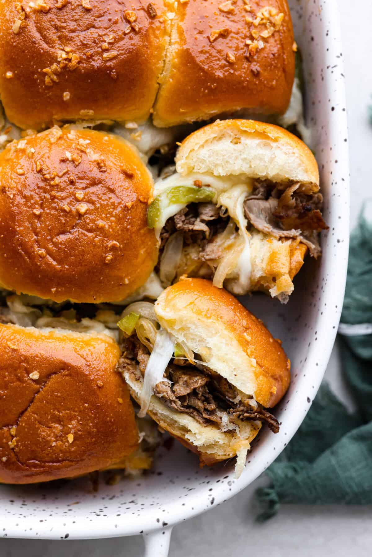 Philly cheesesteak sliders are a fun and delicious twist on a classic. These Philly cheesesteaks are filled with thinly sliced steak, caramelized onions, crisp peppers, and, of course, melty cheese. Overhead shot of Philly cheesesteak sliders in a baking dish with a couple of sliders turned on their side,
