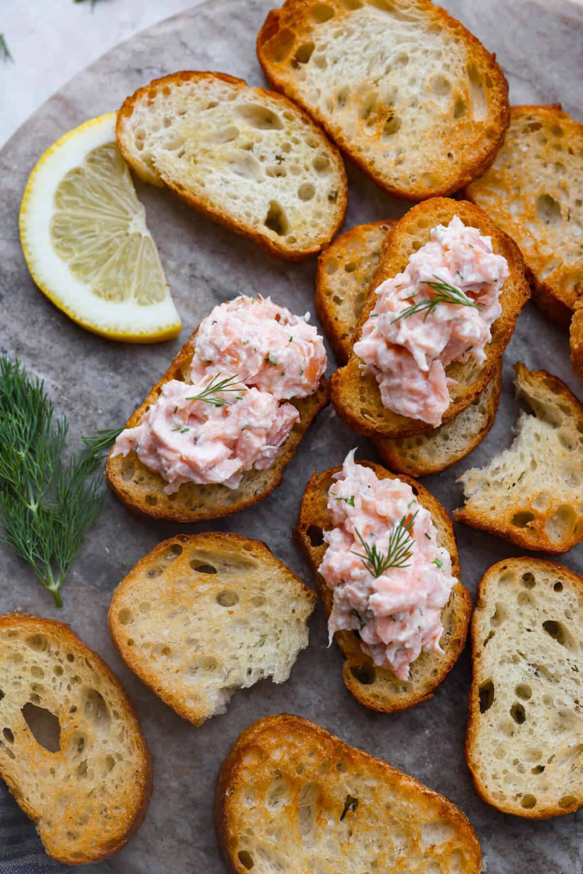 Salmon rillettes is a creamy, flavorful spread made with fresh and smoked salmon blended with lemon, sour cream, and herbs. It is perfect for spreading on crackers or toasted baguette slices. This elegant appetizer is a crowd-pleaser for any occasion! Overhead shot of salmon rillettes on crostini on a serving platter with fresh dill.