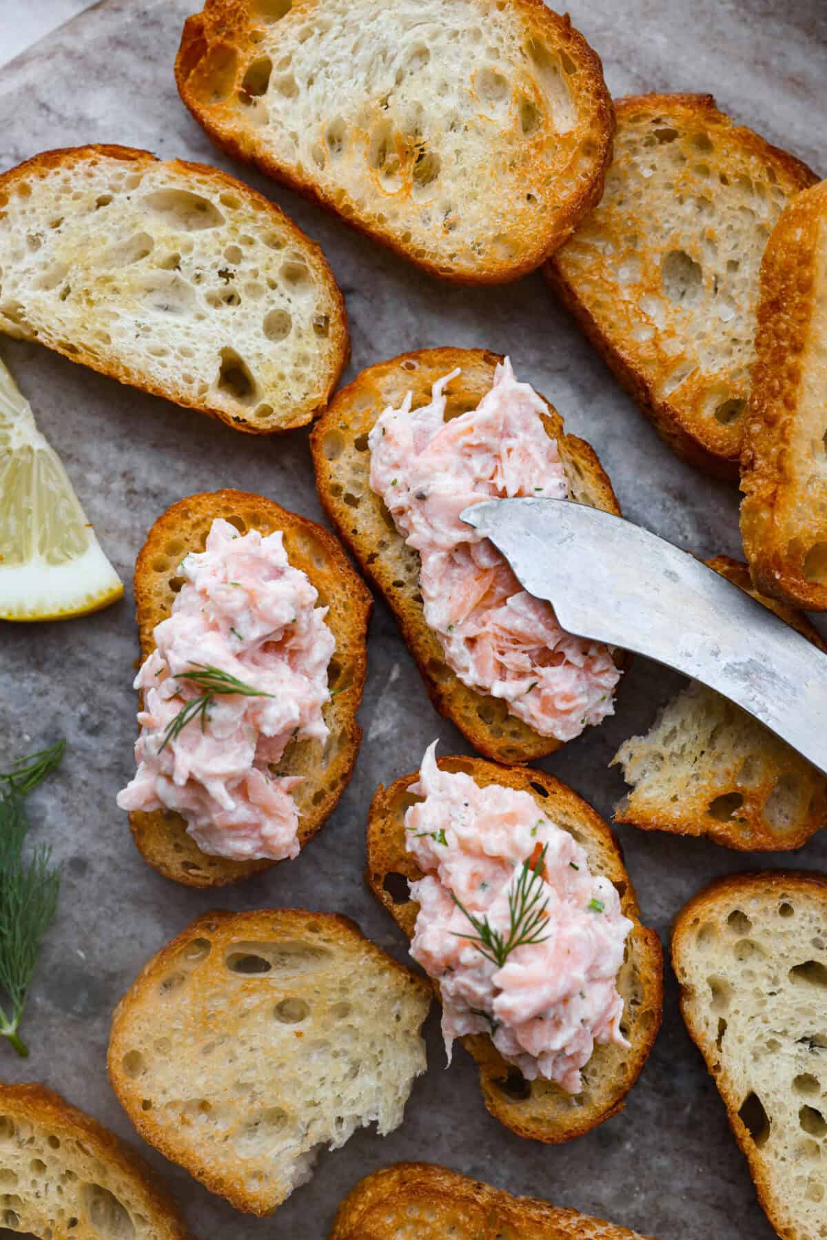 Salmon rillettes is a creamy, flavorful spread made with fresh and smoked salmon blended with lemon, sour cream, and herbs. It is perfect for spreading on crackers or toasted baguette slices. This elegant appetizer is a crowd-pleaser for any occasion! Close up shot of someone spreading salmon rillettes on crostini.