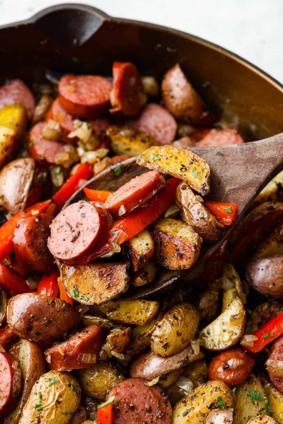 This sausage and potato skillet recipe is meant to be a simple one skillet meal! It has tender baby potatoes, smoked sausage, peppers, and delicious seasonings that take it over the top. It's hearty, satisfying and loaded with flavor! Close view of a wood spatula lifting up sausage, potatoes, and peppers.