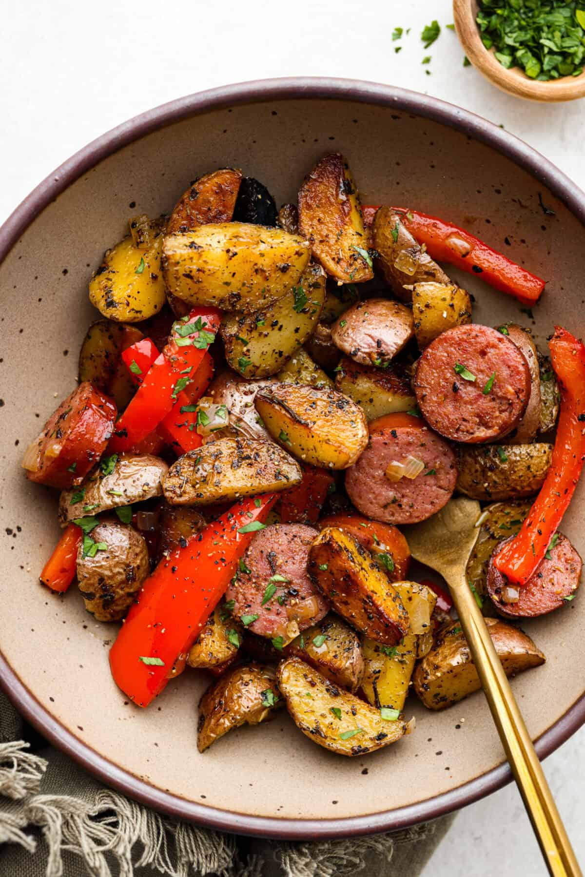 This sausage and potato skillet recipe is meant to be a simple one skillet meal! It has tender baby potatoes, smoked sausage, peppers, and delicious seasonings that take it over the top. It's hearty, satisfying and loaded with flavor! To view of sausage and potato skillet in a bowl with a gold fork.