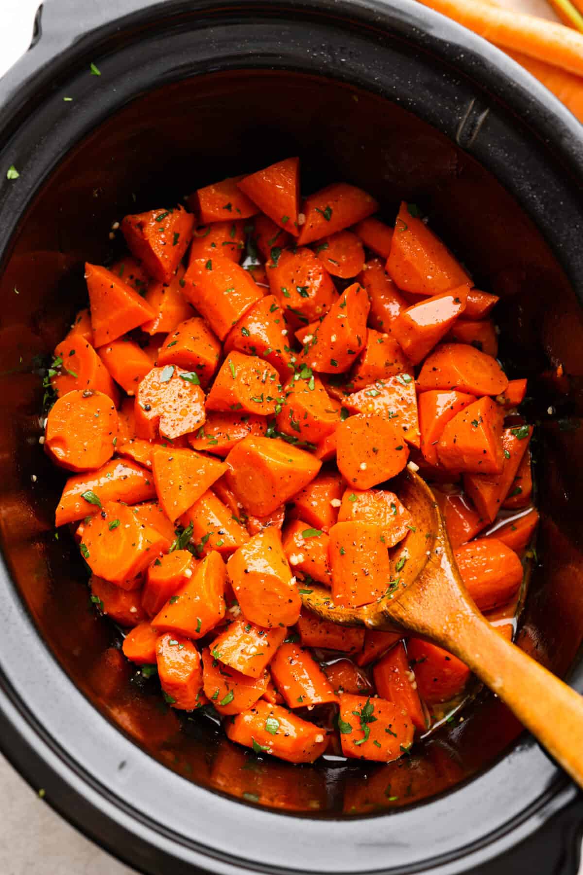 These crockpot carrots will become fork-tender from slowly cooking in your crockpot all day. The carrots have a delicious sweet and savory flavor from the sticky honey and bold garlic, all slathered in butter. They are the perfect side for your next Thanksgiving or Easter feast. Overhead shot of crockpot carrots in a crockpot with a wooden serving spoon.