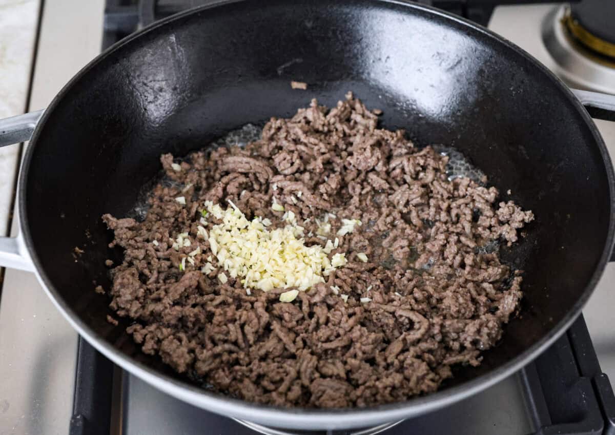 Mongolian Ground Beef and Noodles is a flavorful dinner that’s ready in 30 minutes! This dish combines tender noodles, savory ground beef, and a sweet and tangy Asian-inspired sauce for a meal that’s as comforting as it is delicious! Angle shot of cooked ground beef in a skillet with minced garlic added on top.