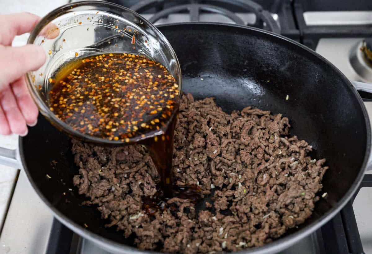 Mongolian Ground Beef and Noodles is a flavorful dinner that’s ready in 30 minutes! This dish combines tender noodles, savory ground beef, and a sweet and tangy Asian-inspired sauce for a meal that’s as comforting as it is delicious! Angle shot of someone pouring the sauce of the drained beef and garlic.