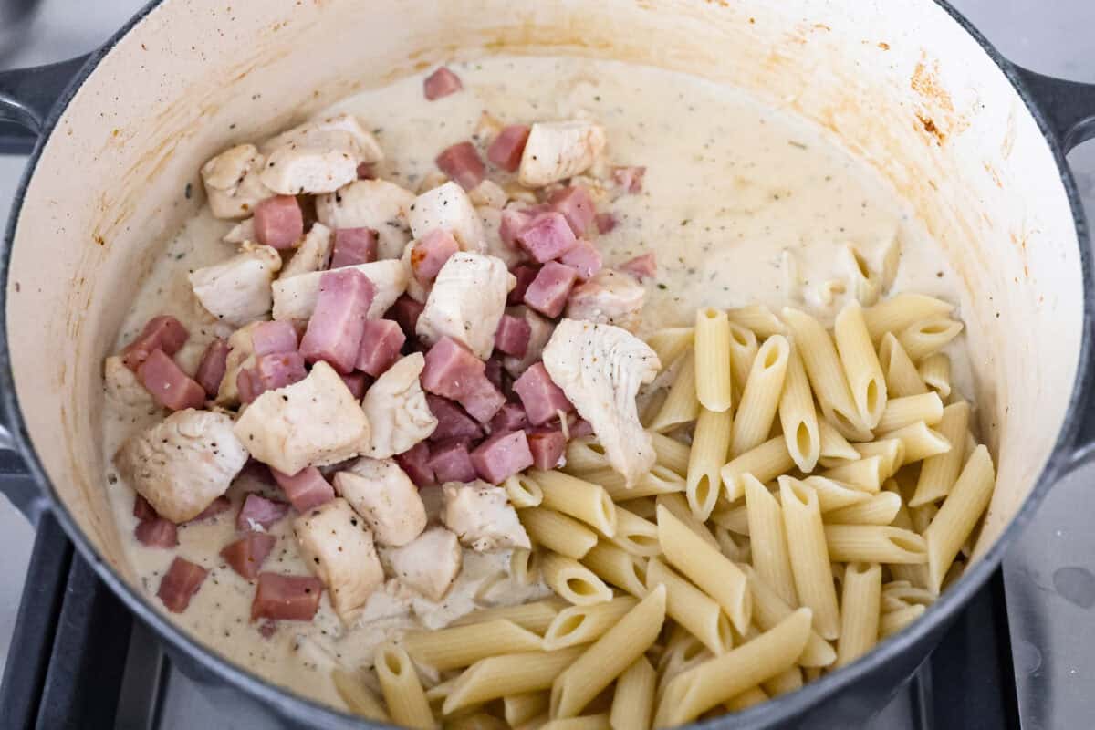 Chicken cordon bleu pasta is a creamy, flavorful dish that's a fun twist on the classic. It combines savory ham, tender chicken, melty cheese, and delicious seasonings. Easy to make and sure to please! Angle shot of the ham, chicken and noodles added to the seasoned sauce in the pot.