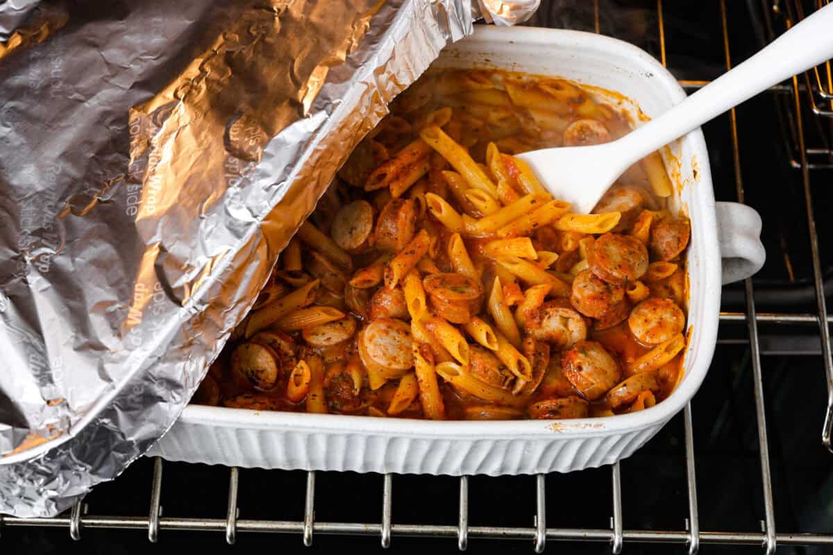 This one-pan Italian Sausage Casserole is the ultimate comfort food! With tender pasta, flavorful sausage, rich marinara, and gooey melted cheese, it’s an easy dinner that’s perfect for busy nights. It's a recipe the whole family will love! Angle shot of someone lifting the tin foil off the pan and stirring the noodle bake.