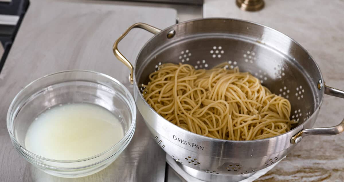 Spaghetti Aglio e Olio uses simple ingredients to create a dish bursting with flavor! With just garlic, olive oil, chili flakes, pasta, and parmesan cheese, this classic Italian recipe is a go-to for a tasty meal in less than 30 minutes. Angle shot of cooked spaghetti noodles in a colander with pasta water next to it in a separate bowl.