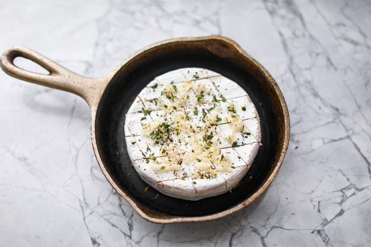 Warm and gooey baked brie is a hit for any occasion! With just five simple ingredients and less than 20 minutes, you will have a rich, creamy appetizer that's impossible to resist. Scoop it up with your favorite crackers or bread! Overhead shot of the circle cheese with garlic and herbs sprinkled over the top of in a skillet.