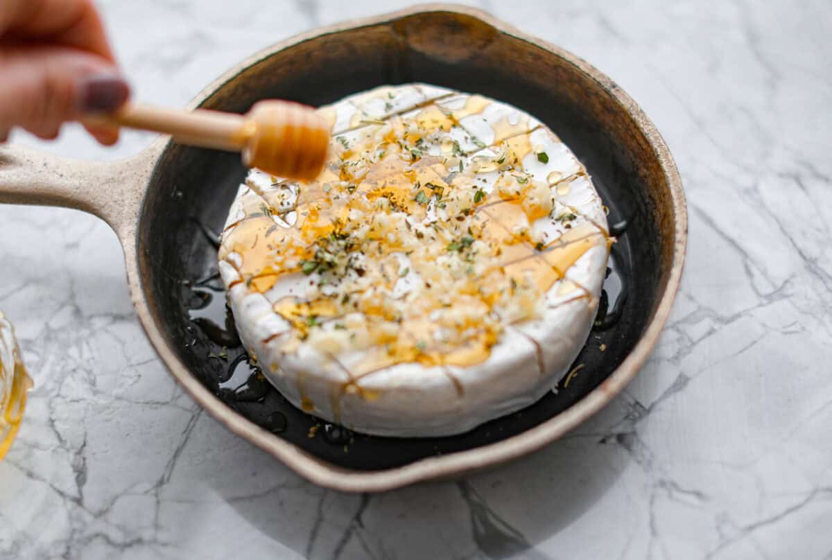 Warm and gooey baked brie is a hit for any occasion! With just five simple ingredients and less than 20 minutes, you will have a rich, creamy appetizer that's impossible to resist. Scoop it up with your favorite crackers or bread! Angle shot of someone drizzling honey over garlic and herbs on the circle of cheese in a skillet.