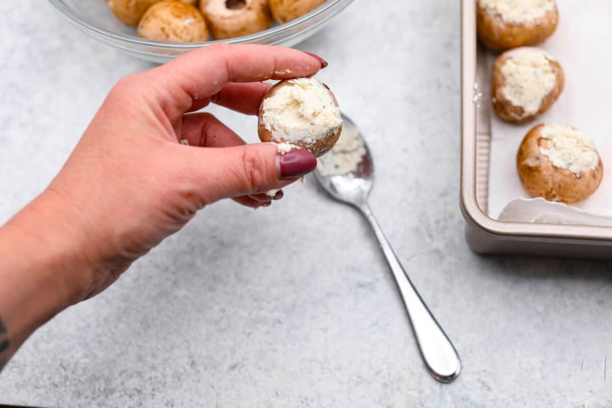 Boursin stuffed mushrooms are a fun, delicious appetizer or side dish. These creamy, cheesy stuffed mushrooms are packed with flavorful Boursin cheese and topped with a delightful breadcrumb crunch. Overhead shot of someone holding a mushroom with Boursin cheese stuffed into the opening.