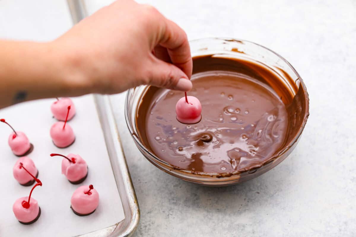 Homemade cherry cordials are a tasty, nostalgic treat! They're made with sweet cherries that are coated in a rich chocolate shell. Make them for Christmas or Valentineโs Day, and watch them disappear! Angle shot of someone dipping fondant dipped cherries into the chocolate.