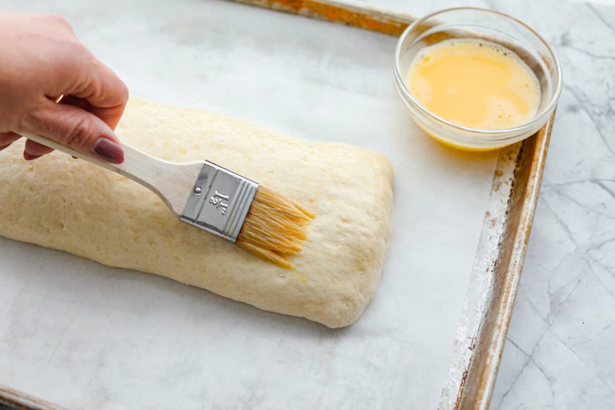 This Breakfast Stromboli is the best morning treat! It's packed with fluffy eggs, savory sausage, crispy bacon, and melty cheese, all wrapped in golden pizza dough. Perfect for holiday brunches or everyday family breakfasts! Angle shot of someone spreading the gg wash on the Stromboli.