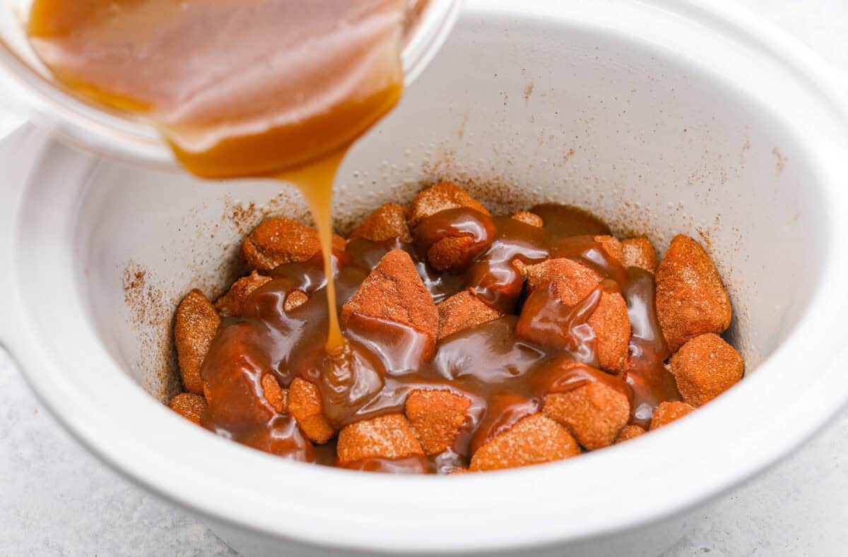 Crockpot monkey bread is an easy, gooey treat made in your slow cooker. It’s packed with soft biscuit pieces, cinnamon-sugar flavor, and a sweet buttery glaze. No oven needed! Angle shot of someone pouring brown sugar and butter glaze over the top of the biscuits.