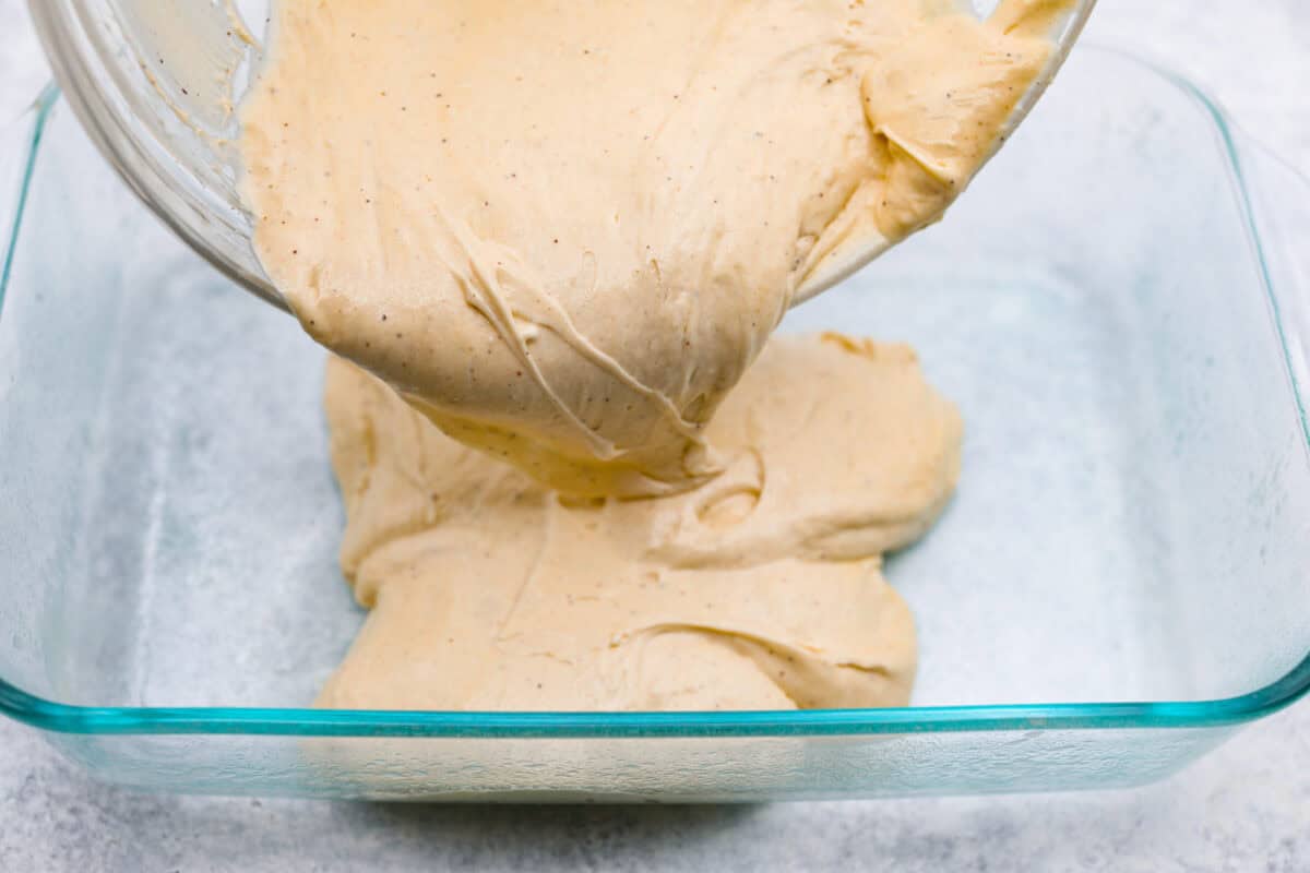 Eggnog Poke Cake could not be more perfect for the holiday season! It combines the rich flavor of eggnog with the soft, moist texture of a classic poke cake. Add fluffy whipped topping and a sprinkle of nutmeg and cinnamon to really wow your family! Angle shot of someone pouring the cake batter into a prepared baking dish.