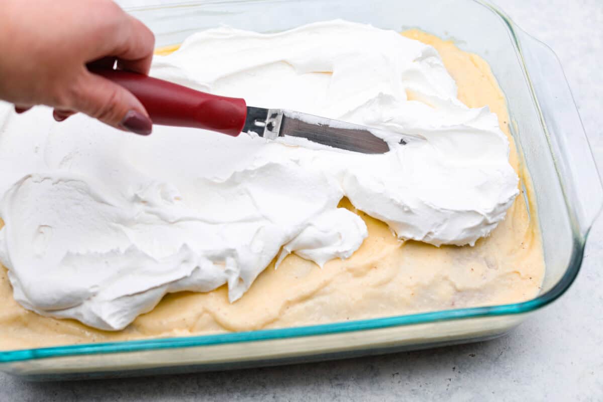 Eggnog Poke Cake could not be more perfect for the holiday season! It combines the rich flavor of eggnog with the soft, moist texture of a classic poke cake. Add fluffy whipped topping and a sprinkle of nutmeg and cinnamon to really wow your family! Angle shot of someone spreading whipped topping over the spread topping.