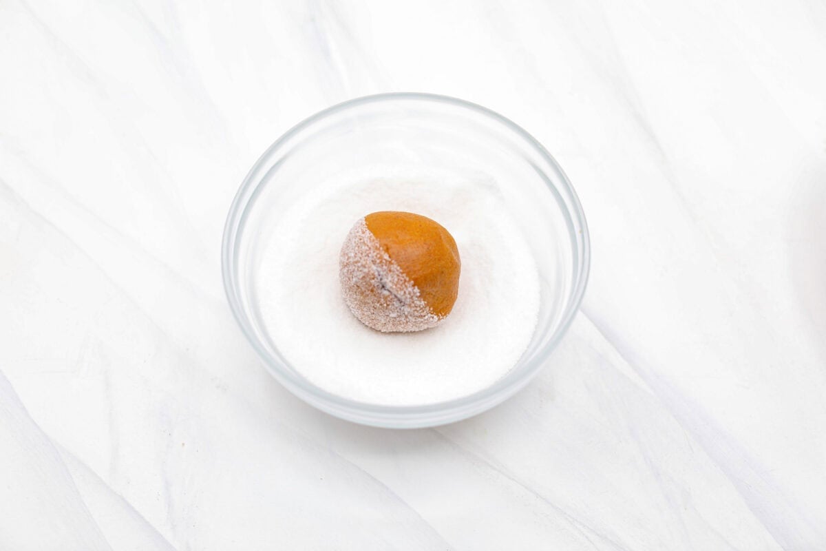 These gingerbread crinkle cookies are incredibly soft and flavorful! Their deep molasses flavor, spiced to perfection and rolled in powdered sugar, is so good with a glass of milk. They are the perfect cookies to make for Santa this holiday season. Angle shot of gingerbread crinkle cookie dough ball in a bowl of granulated sugar.