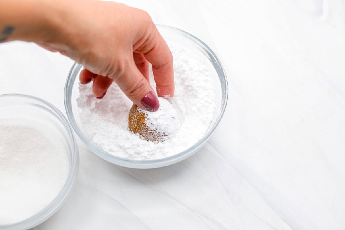 These gingerbread crinkle cookies are incredibly soft and flavorful! Their deep molasses flavor, spiced to perfection and rolled in powdered sugar, is so good with a glass of milk. They are the perfect cookies to make for Santa this holiday season. Angle shot of someone rolling a dough ball in powdered sugar.