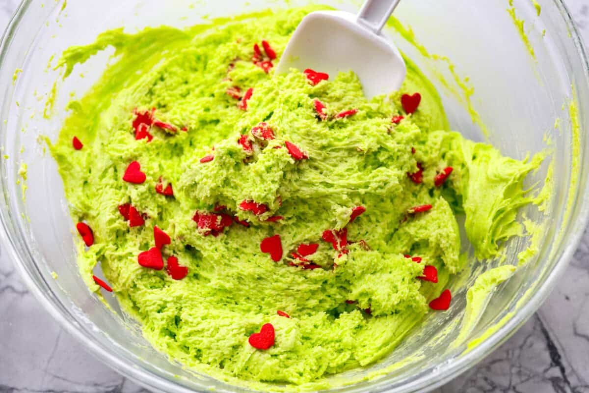 Grinch dip is a sweet and scrumptious dip perfect for any holiday party or gathering. It combines delicious cake mix, fluffy whipped topping, and smooth yogurt or pudding to create a fun dip for cookies, treats, or fruit. Overhead shot of someone folding in heart shaped sprinkles into the dip.