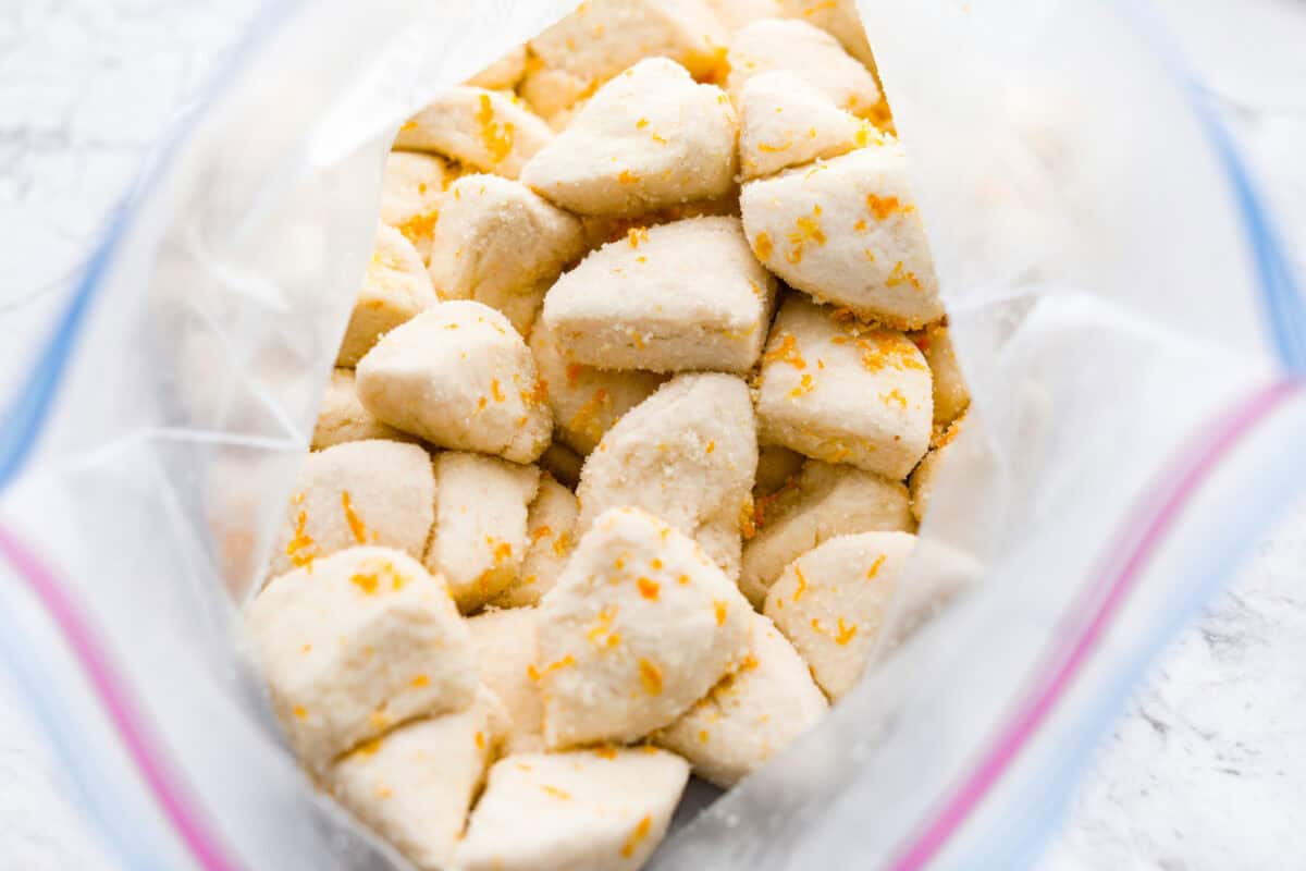 Orange monkey bread is a fun twist on a classic sticky pull-apart bread. It's packed with soft, buttery biscuit pieces, bursts of citrusy orange zest, and a sticky, sweet glaze. Everyone will want to get their hands on this delicious dish! Overhead shot of biscuit quarters in a ziplock back mixed with sugar and orange zest.