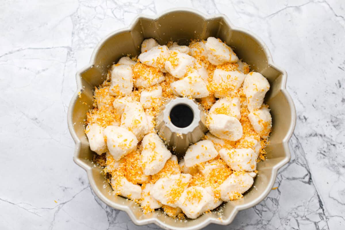 Orange monkey bread is a fun twist on a classic sticky pull-apart bread. It's packed with soft, buttery biscuit pieces, bursts of citrusy orange zest, and a sticky, sweet glaze. Everyone will want to get their hands on this delicious dish! Overhead shot of biscuit pieces and sugar mixture in a greased bundt pan.