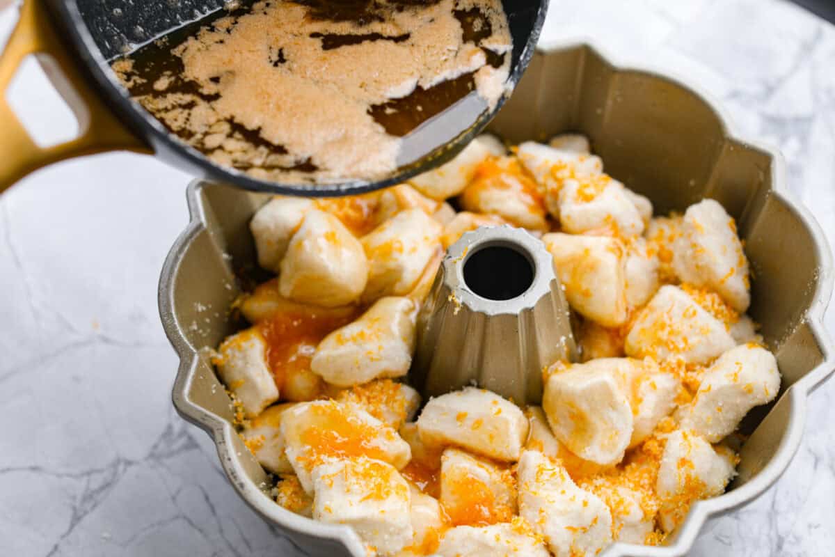 Orange monkey bread is a fun twist on a classic sticky pull-apart bread. It's packed with soft, buttery biscuit pieces, bursts of citrusy orange zest, and a sticky, sweet glaze. Everyone will want to get their hands on this delicious dish! Angle shot of someone pouring the butter and brown sugar sauce over the biscuit and orange zest in the bundt pan.