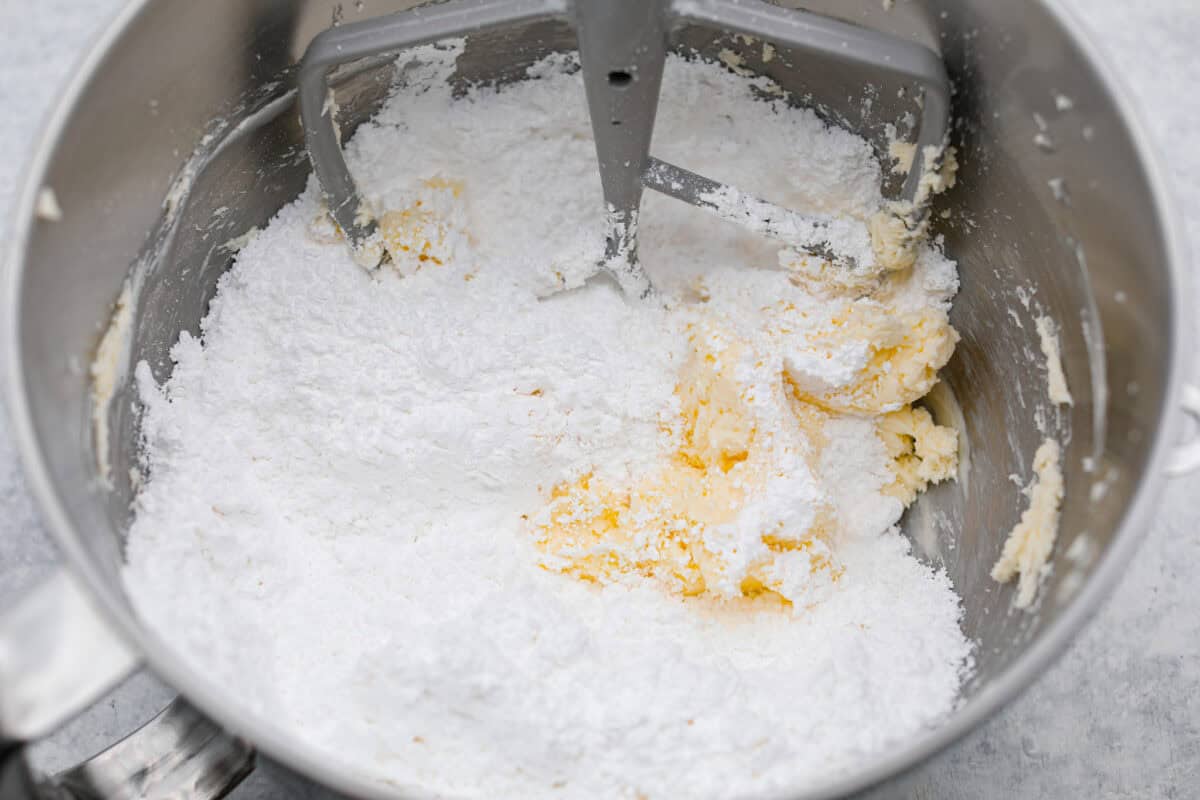 Tom Cruise coconut cake is a dense pound cake with coconut flavor and silky white chocolate throughout. The indulgent frosting is a coconut cream cheese buttercream with sweetened coconut on the outside. It's a coconut lover's dream—but a super sweet one. Overhead shot of powdered sugar dumped in with the butter and cream cheese mixture.