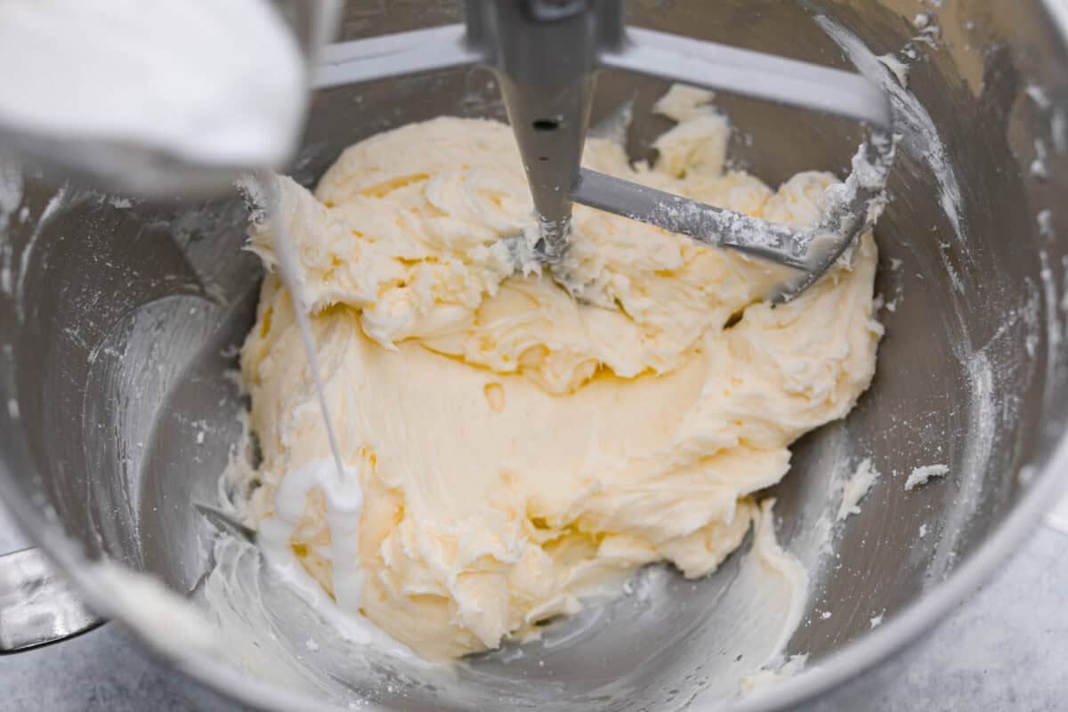 Tom Cruise coconut cake is a dense pound cake with coconut flavor and silky white chocolate throughout. The indulgent frosting is a coconut cream cheese buttercream with sweetened coconut on the outside. It's a coconut lover's dream—but a super sweet one. Overhead shot of someone pouring milk into the frosting mixture.
