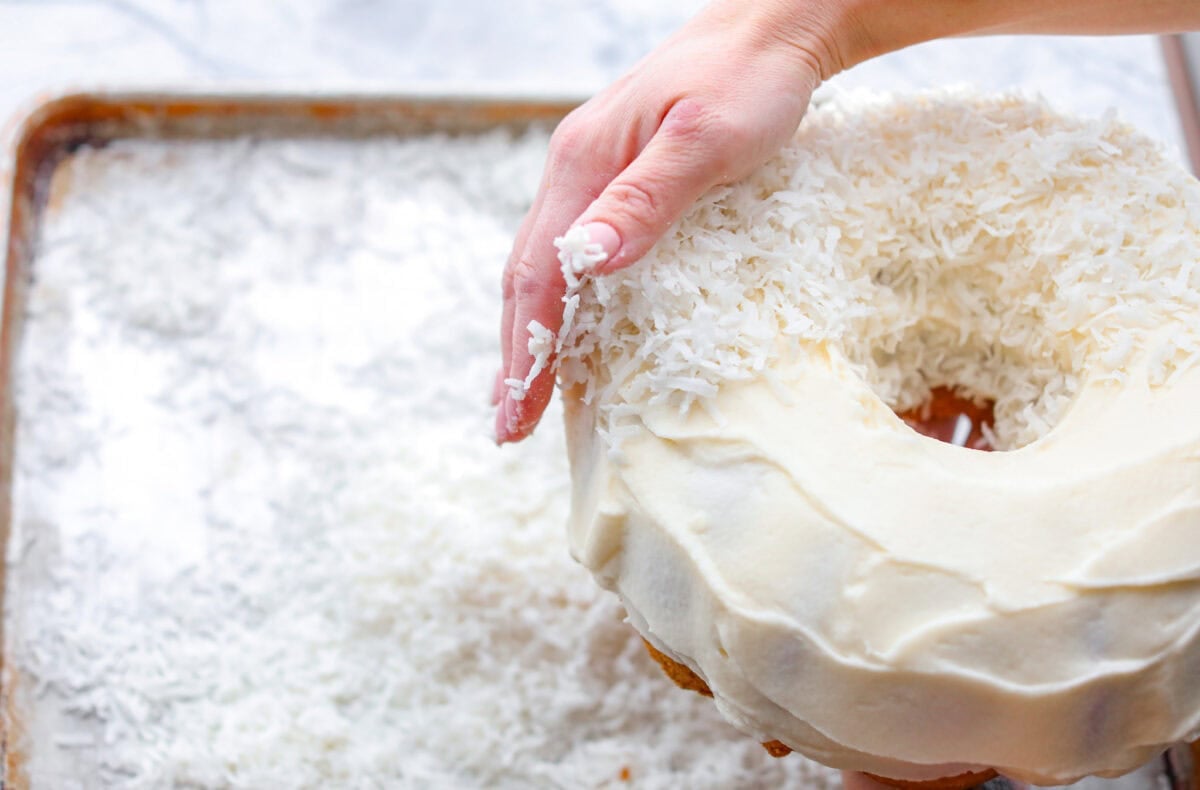 Tom Cruise coconut cake is a dense pound cake with coconut flavor and silky white chocolate throughout. The indulgent frosting is a coconut cream cheese buttercream with sweetened coconut on the outside. It's a coconut lover's dream—but a super sweet one. Angle shot of someone pressing shredded coconut flakes onto the frosted cake.