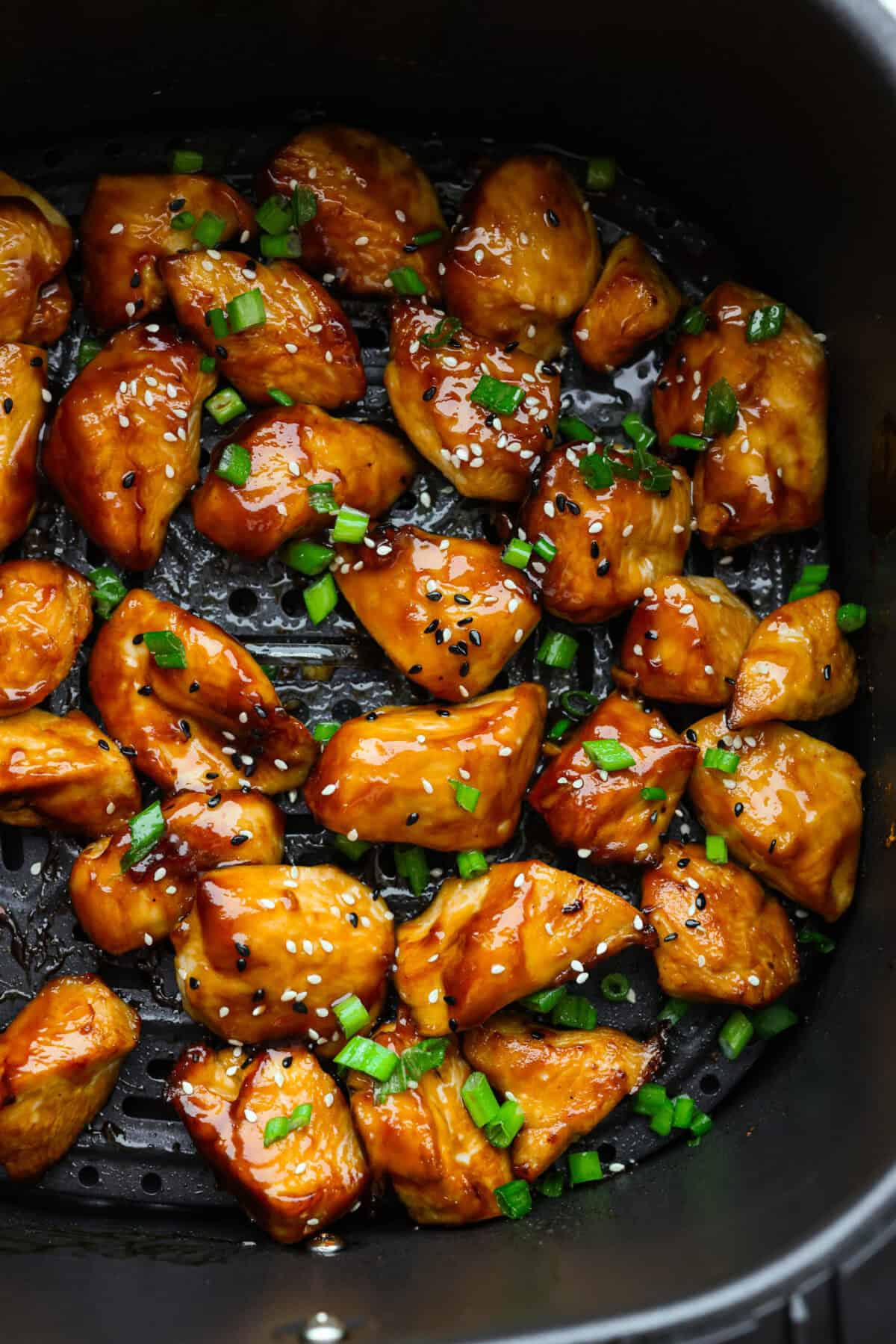 Super flavorful Air Fryer Teriyaki Chicken cooks up perfectly every time! It's tender and juicy on the inside, with a tasty teriyaki sauce coating the outside. Pair with rice and veggies for an easy meal on busy nights! Close up shot of air fryer teriyaki chicken in the bottom of the air fryer basket with sesame seeds and green onions.