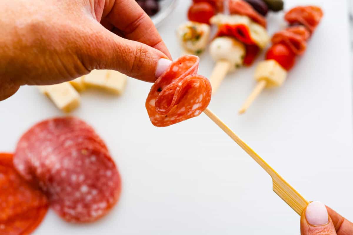 Bring this antipasto charcuterie wreath to your next holiday party! Skewered cheese, deli meats, olives, and pickles shaped into a cute Christmas wreath. A perfect festive appetizer to impress your guests! First photo of the pepperoni being placed on a skewer.
