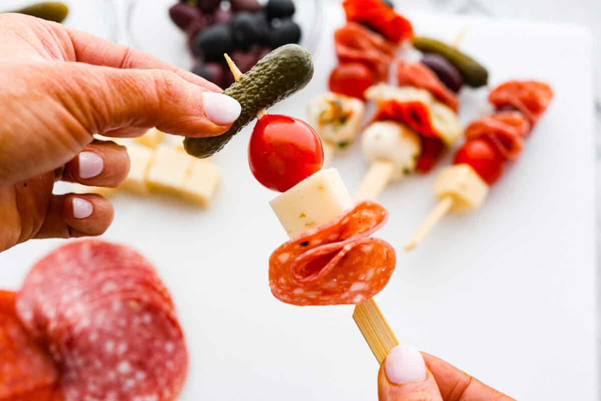 Bring this antipasto charcuterie wreath to your next holiday party! Skewered cheese, deli meats, olives, and pickles shaped into a cute Christmas wreath. A perfect festive appetizer to impress your guests! Second photo of a pickle going on a skewer full of tomato, cheese, and pepperoni.