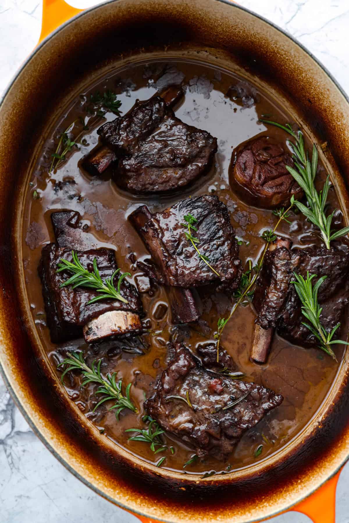Tender braised short ribs will fall off the bone and leave you speechless! Seared then cooked in a red wine reduction, beef broth, and fresh herbs. Every bite is tender perfection and will melt in your mouth! Top view of braised short ribs in a dutch oven pot garnished with herbs.