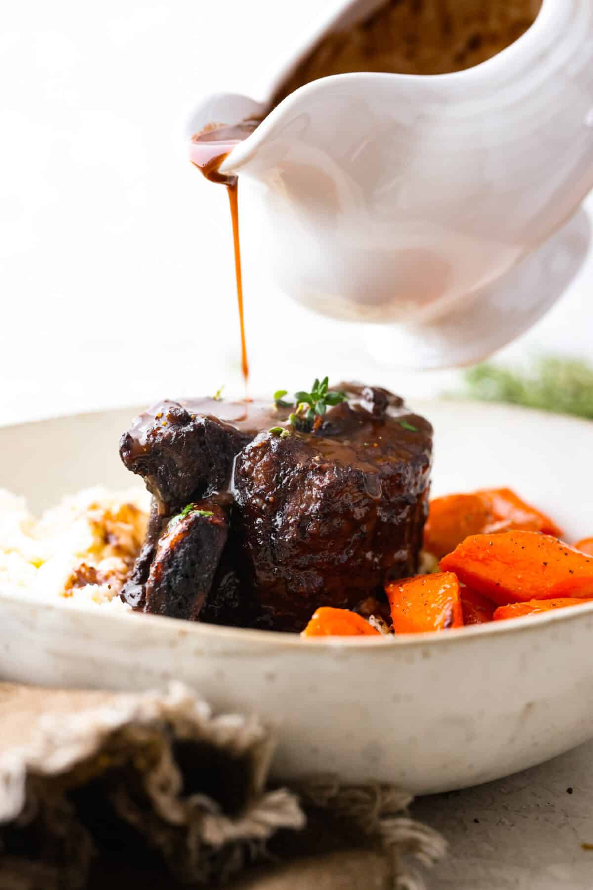 Tender braised short ribs will fall off the bone and leave you speechless! Seared then cooked in a red wine reduction, beef broth, and fresh herbs. Every bite is tender perfection and will melt in your mouth! Close view of the gravy pouring over beef short ribs.
