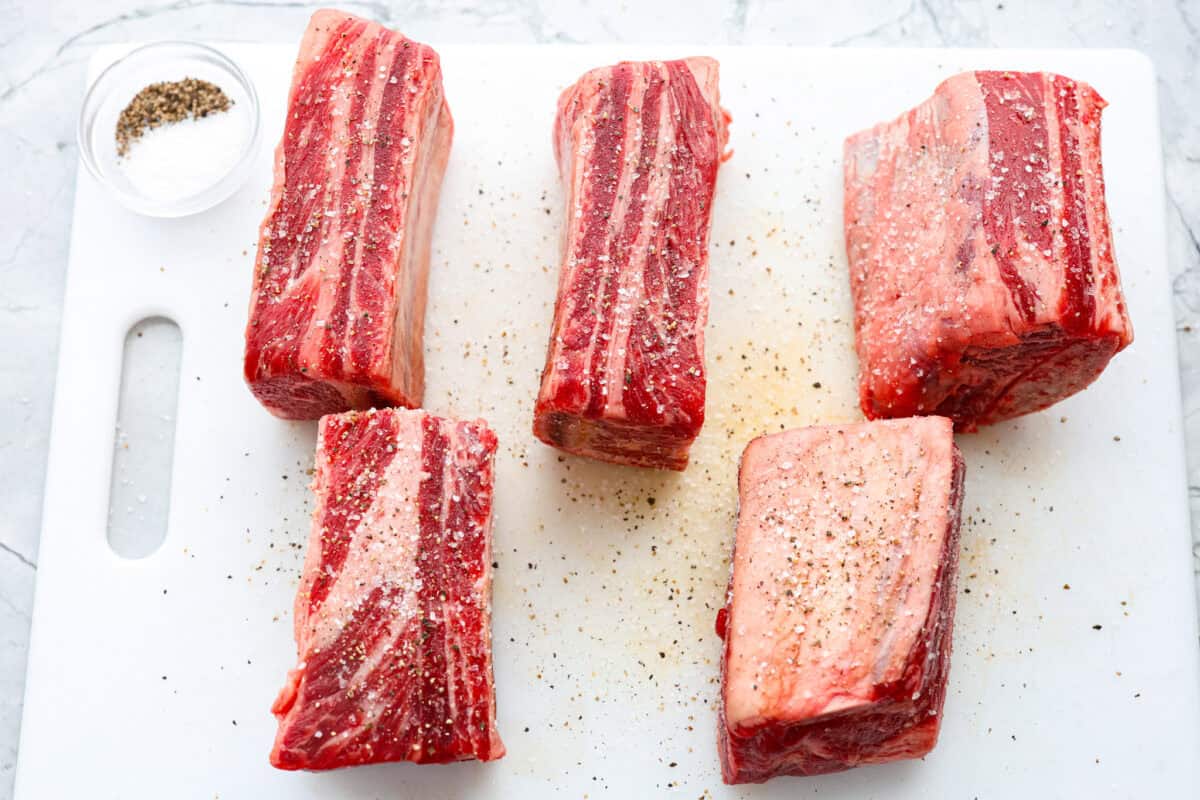 Tender braised short ribs will fall off the bone and leave you speechless! Seared then cooked in a red wine reduction, beef broth, and fresh herbs. Every bite is tender perfection and will melt in your mouth! First photo of the short ribs seasoned with salt and pepper.