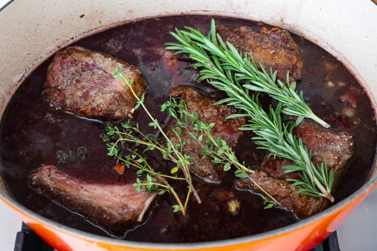 Tender braised short ribs will fall off the bone and leave you speechless! Seared then cooked in a red wine reduction, beef broth, and fresh herbs. Every bite is tender perfection and will melt in your mouth! Sixth photo of the short ribs added back to the pot with fresh herbs.