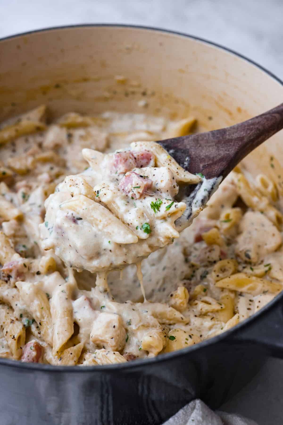 Chicken cordon bleu pasta is a creamy, flavorful dish that's a fun twist on the classic. It combines savory ham, tender chicken, melty cheese, and delicious seasonings. Easy to make and sure to please! Close up shot of a scoop of chicken cordon bleu in a wooden spoon.