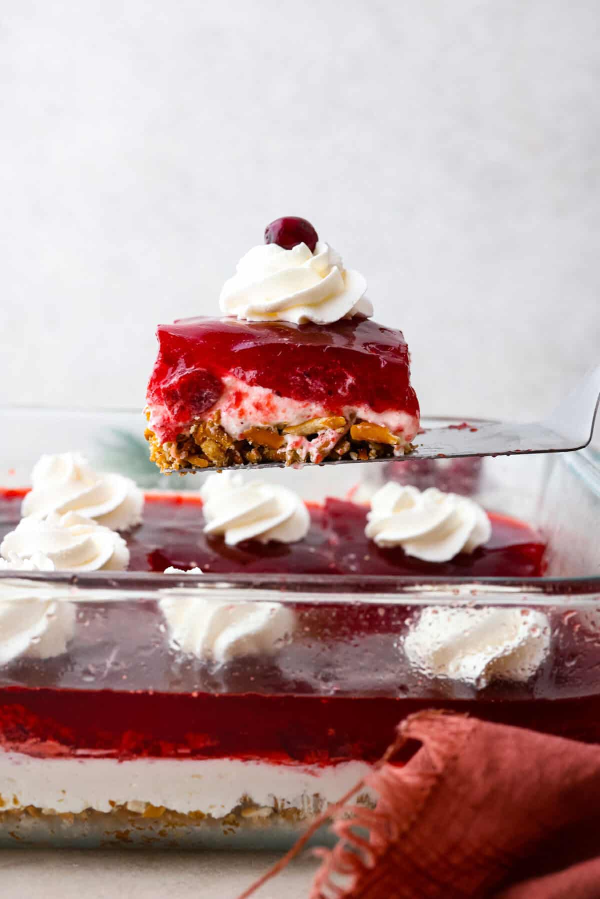 Make this festive cranberry pretzel salad for the holidays! The salty pretzel crust is the perfect balance to the sweet and creamy layers. Cranberry sauce gives it a pop of tartness and makes it holiday-ready! Close side view of a spatula lifting a slice of cranberry pretzel salad from the baking dish.