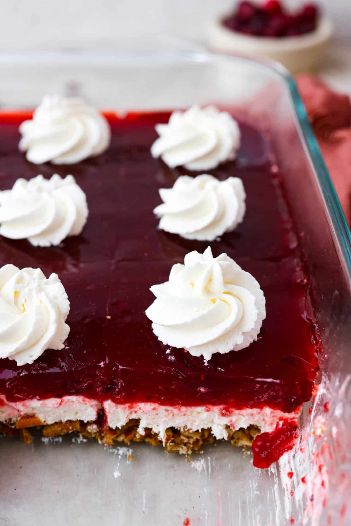 Make this festive cranberry pretzel salad for the holidays! The salty pretzel crust is the perfect balance to the sweet and creamy layers. Cranberry sauce gives it a pop of tartness and makes it holiday-ready! Close top view of cranberry pretzel salad in a baking dish with dollops of whipped cream on top.